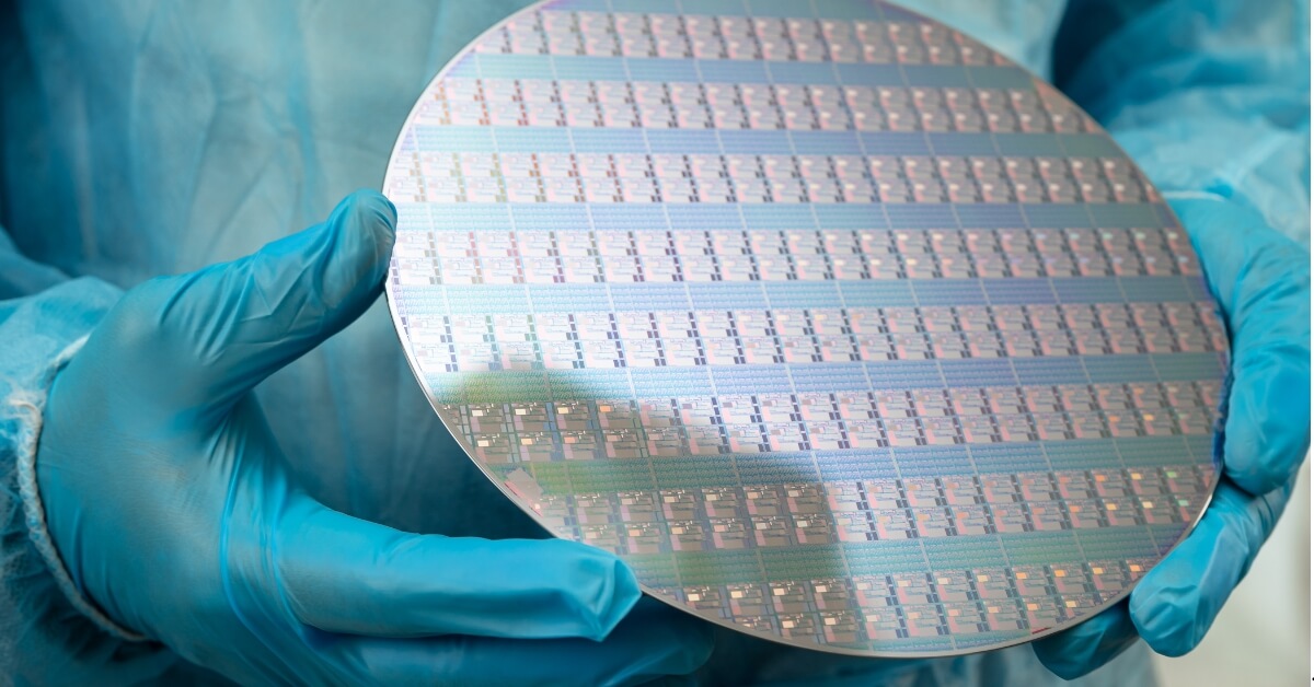 A gloved hand holds a silicon wafer covered in a grid of small, rectangular integrated circuits.