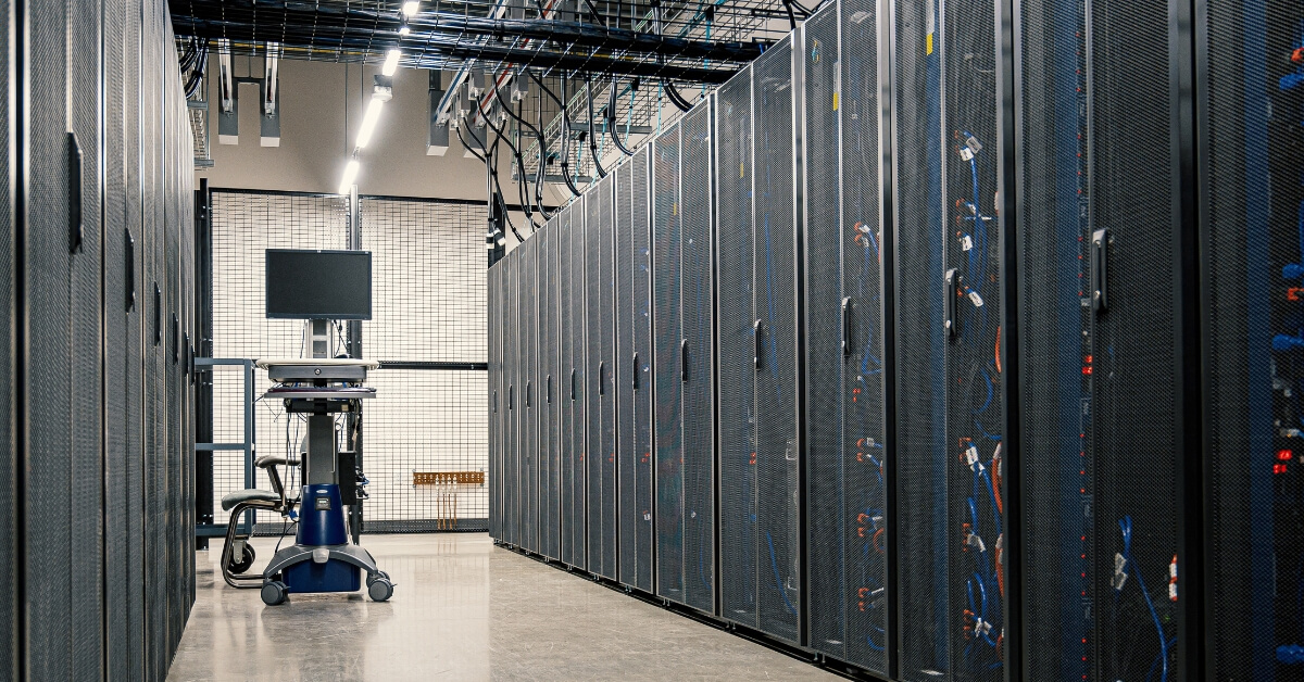 A server room with rows of black server racks and a mobile monitor cart in the aisle.