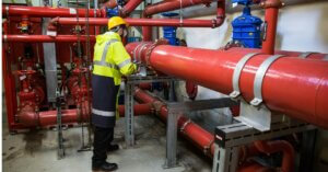 A person in a yellow hard hat and high-visibility vest works on a system of fire protection pipes.