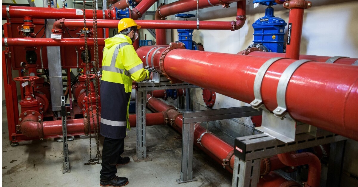 A person in a yellow hard hat and high-visibility vest works on a system of fire protection pipes.